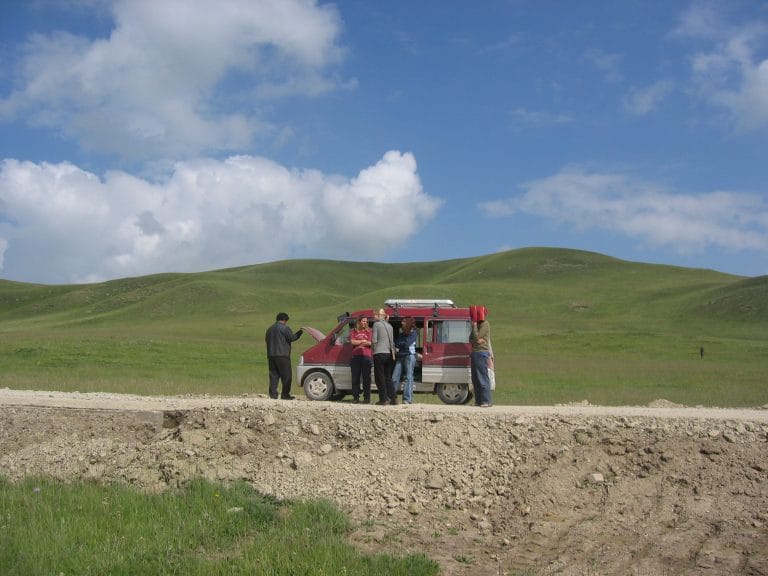 Groep reizigers bij een rode minibus op het groene Ruoergai-plateau in Sichuan, blauwe lucht met wolken