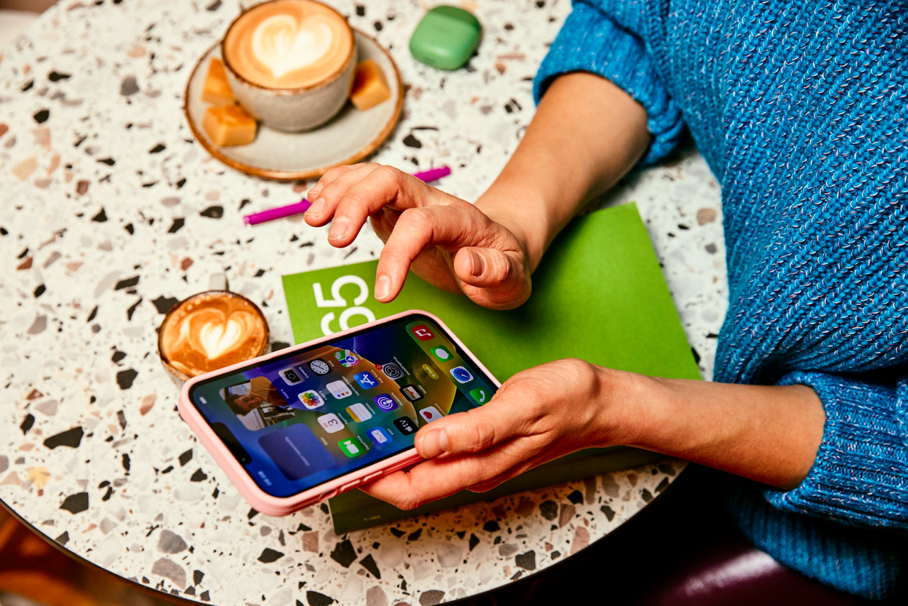 Persoon zit aan tafel met een kop koffie en gebruikt haar mobiele telefoon